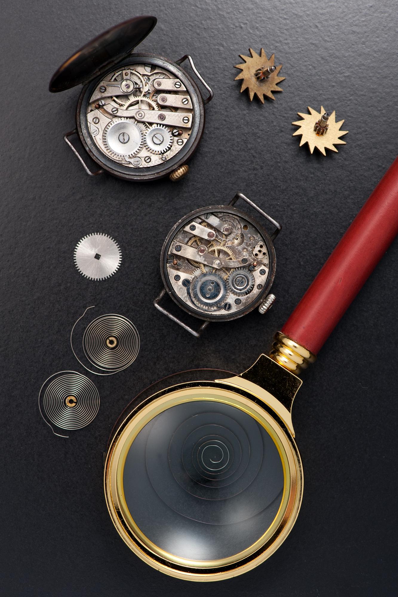 Loupe con manico rosso accanto a due movimenti di orologio da tasca smontati, molle di carica e ruote dentate su sfondo scuro