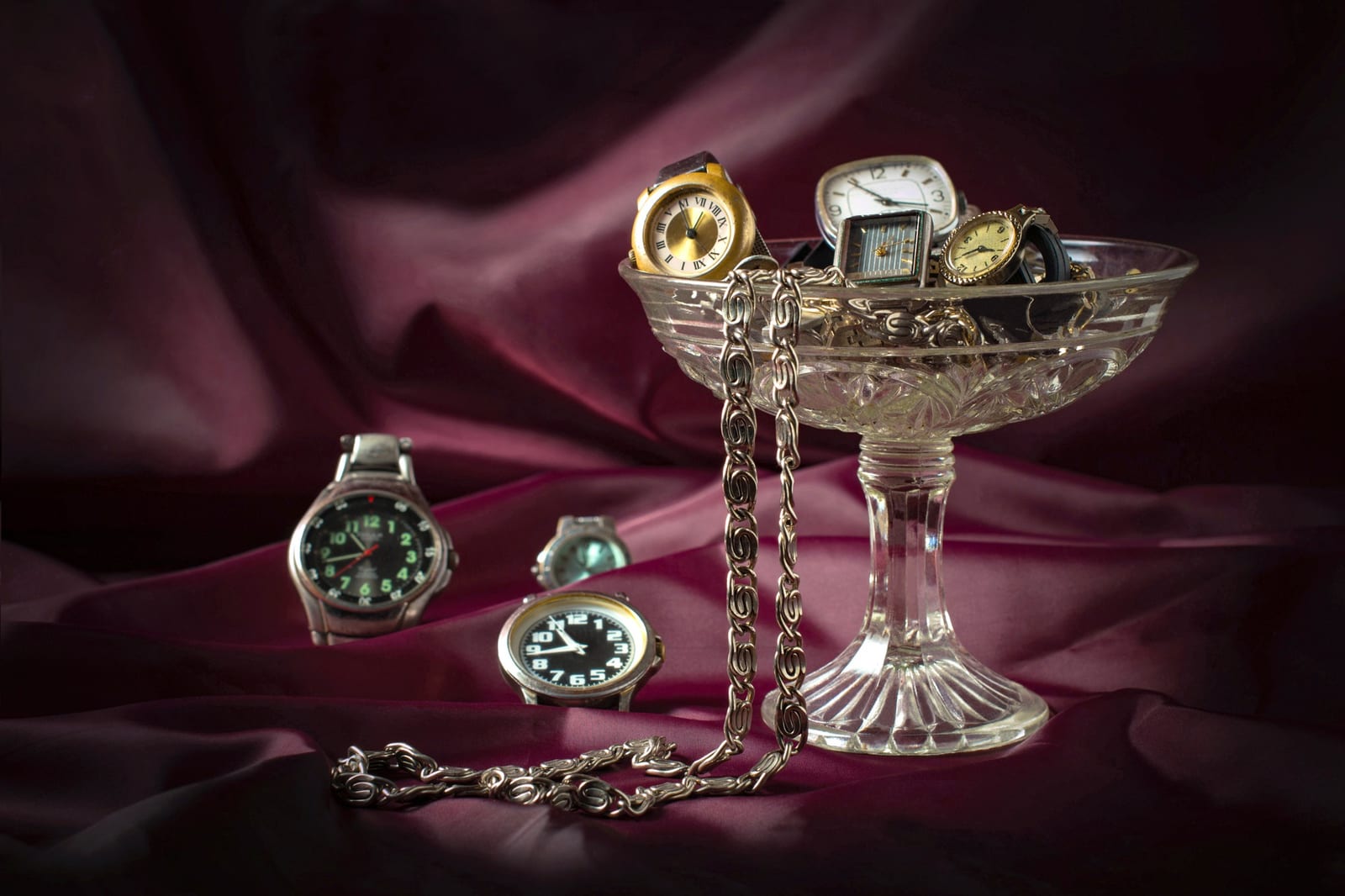 Coppa di cristallo con diversi orologi da polso e da tasca vintage appoggiata su un tessuto bordeaux